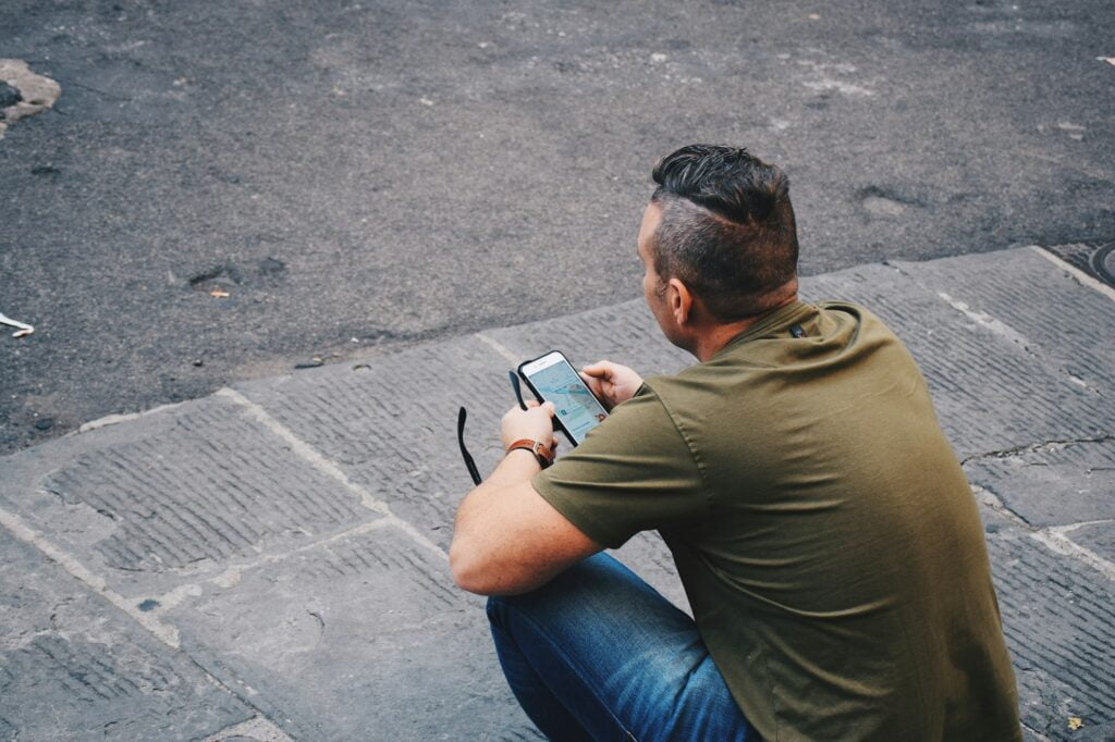 A man is sitting on a sidewalk and looking at a map on his mobile phone